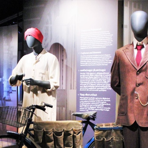 Mannequins dressed in traditional and formal attire next to a bicycle with panniers in a museum display.