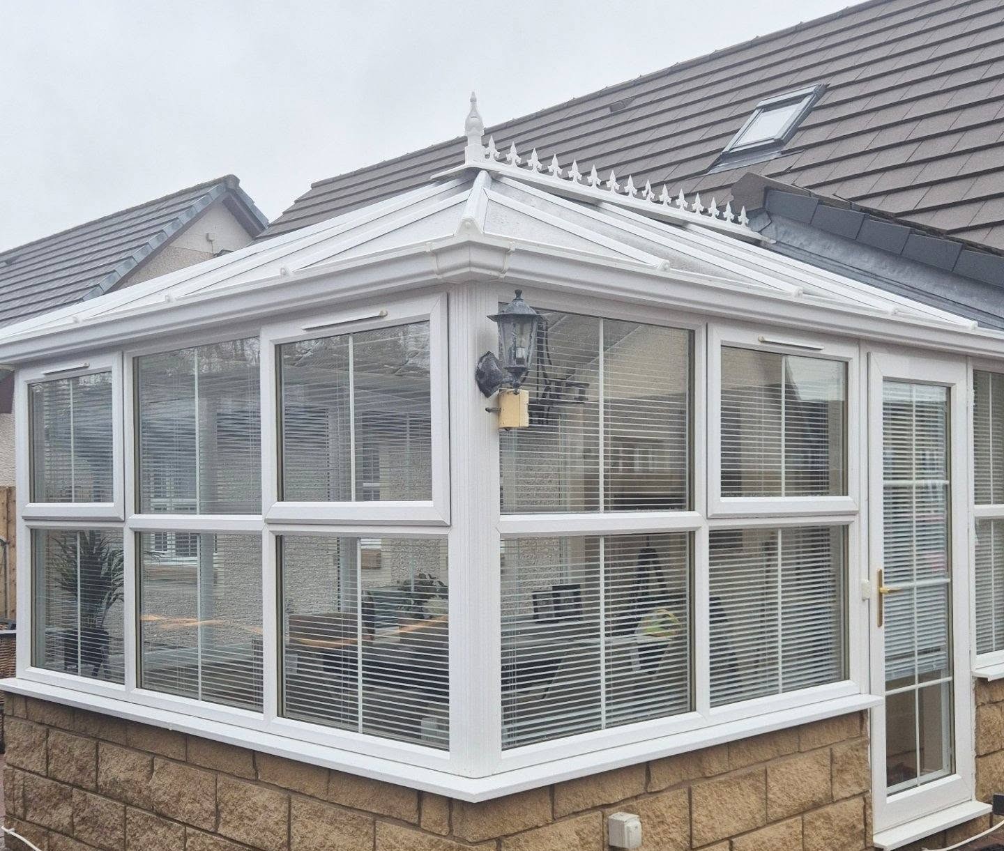 A white conservatory with large glass windows, blinds, a pitched roof, and a brown brick base. It has an ornate roof trim and an outdoor wall lantern.