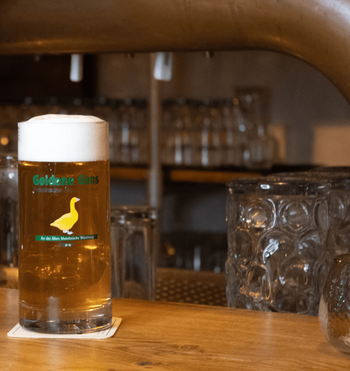 Zwei Biergläser mit dem Logo „Goldene Gans“ stehen auf einer Theke; im Hintergrund leere Gläser und ein Zapfhahn.
