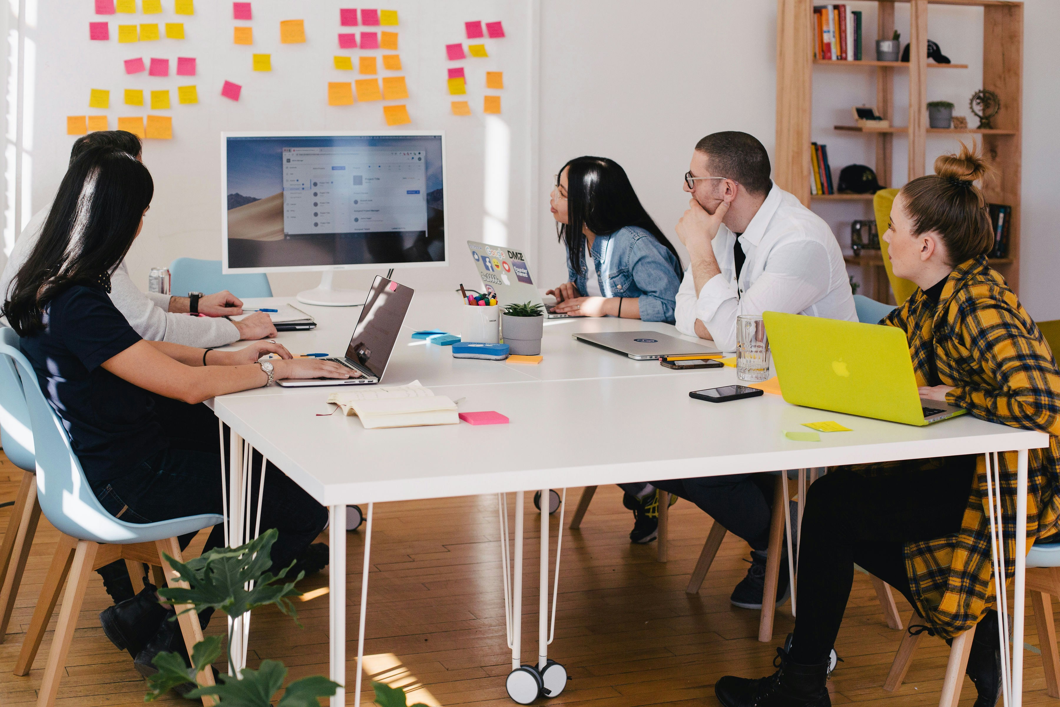 Six  professionals in a modern office setting having a meeting around a white table with laptops and sticky notes in the background.