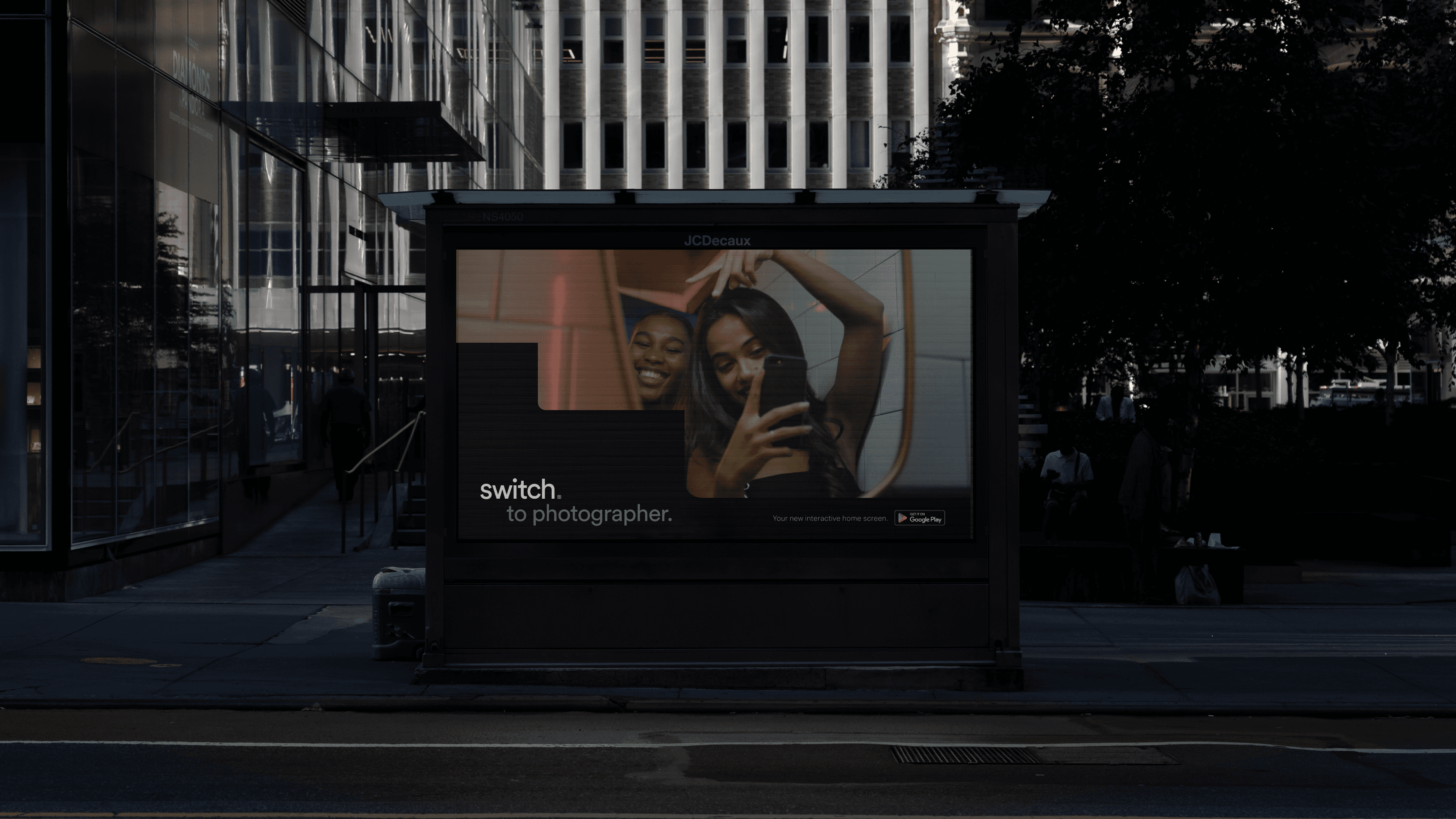 A digital billboard displays a photograph of a woman holding a smartphone, with text reading “switch to photographer.” Below the advertisement, text reads “switch to photographer.” A “Google” logo is visible in the bottom right corner. The billboard is mounted on a dark structure on a sidewalk, with a building and trees in the background.