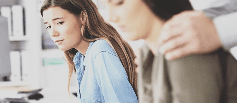 A young woman sitting apart, distressed, while two colleagues comfort each other