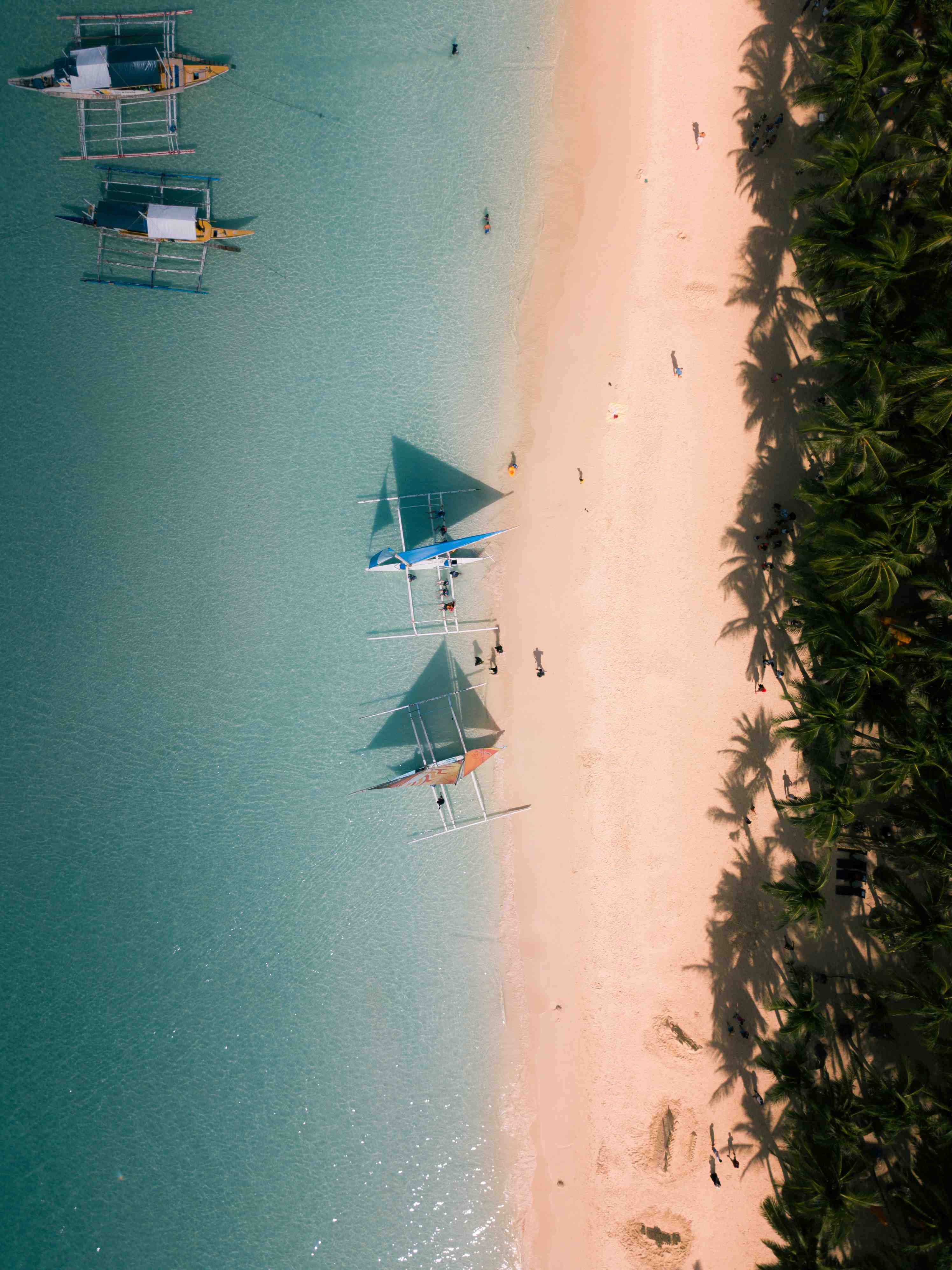 Aerial view of a serene beach in Snaur with turquoise water. Three boats with sails are docked near the shore. Palm trees line the sandy beach, casting long shadows.