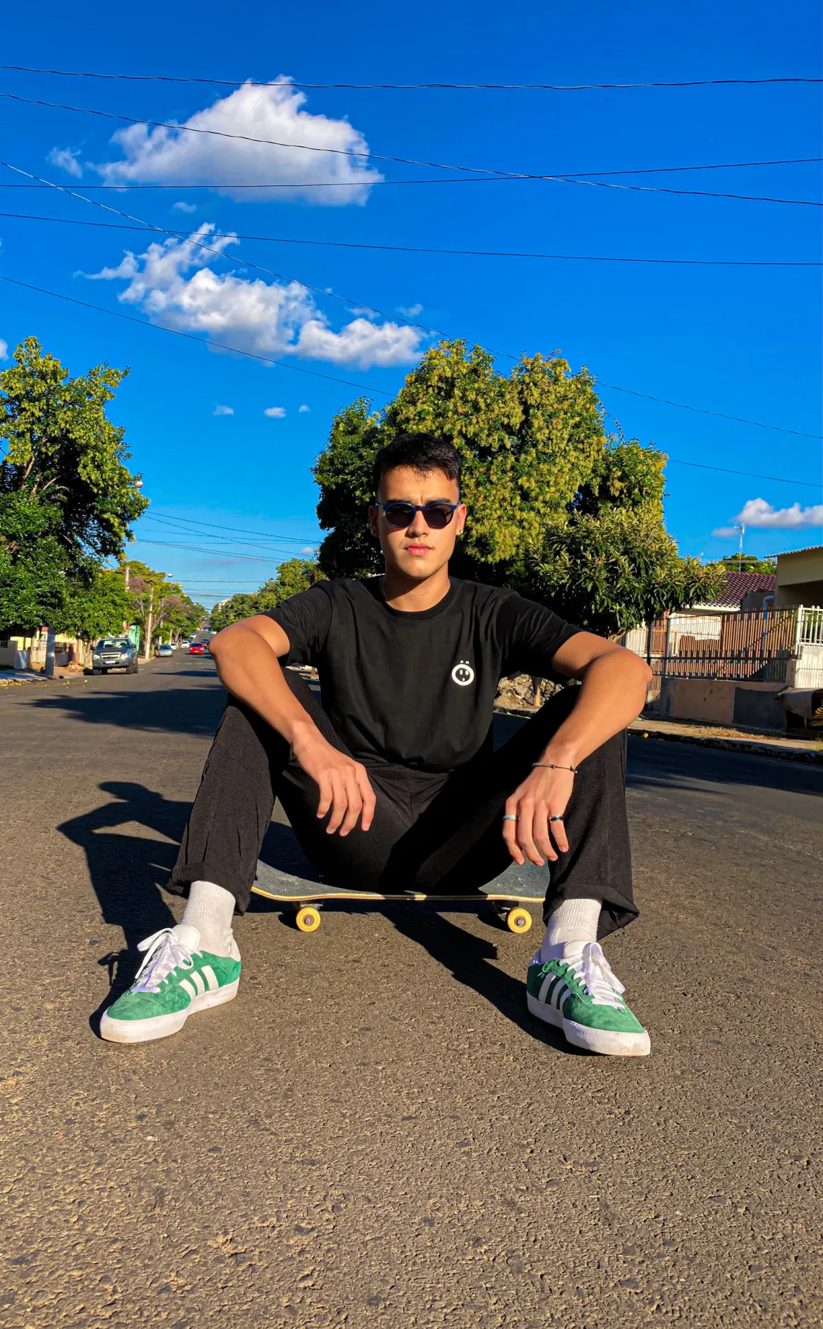 A young man in sunglasses sits on a skateboard in the middle of a sunlit residential street, wearing a black T-shirt with the white surprised-face icon on the left chest.