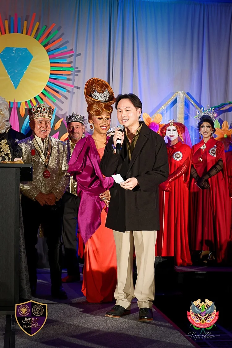 Uma pessoa em traje formal fala ao microfone no palco de um evento com um cenário colorido e festivo, cercada por outras pessoas em fantasias elaboradas e coroas majestosas, refletindo a atmosfera de uma celebração ou cerimônia de premiação.