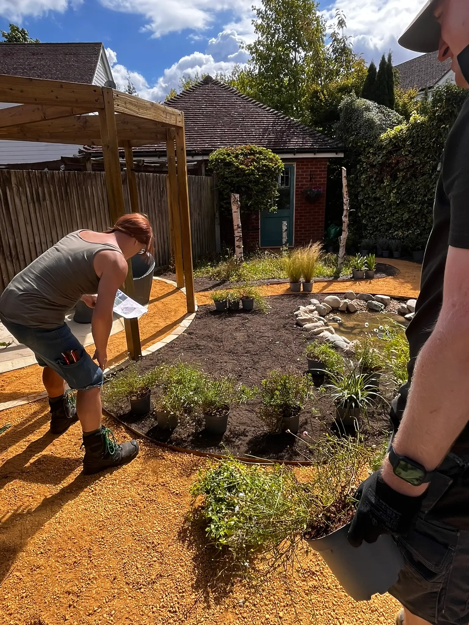 Two people work in a garden, planting shrubs and landscaping under a blue sky with some clouds.