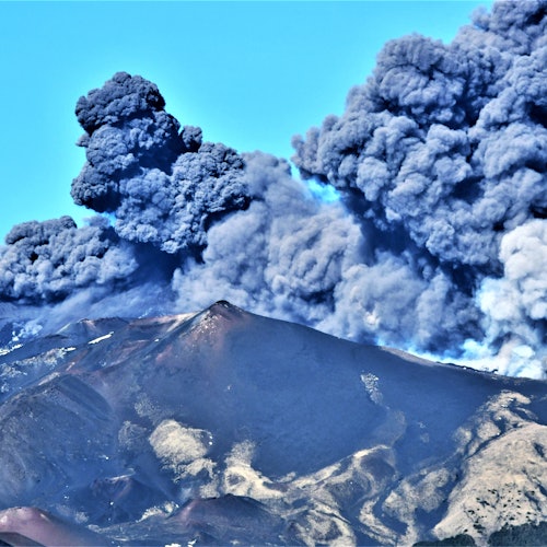 Nuvens densas e escuras de fumaça e cinzas saem de um vulcão em erupção sob um céu claro e azul.