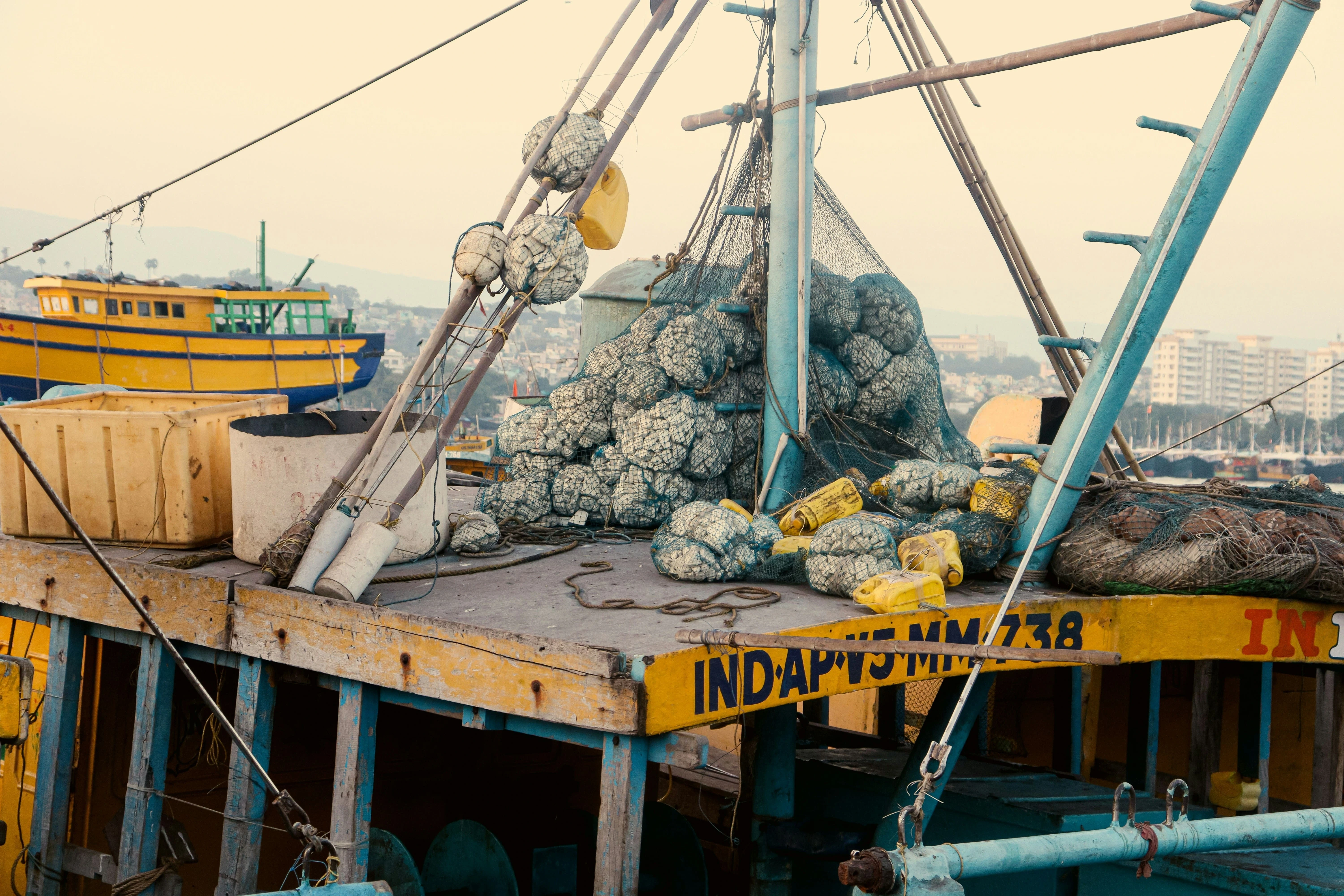 Fishing boat deck loaded with nets and baskets of shellfish, with harbor buildings and other boats in the background.