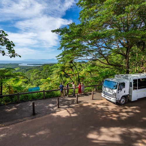 Quattro persone si trovano su un punto panoramico con una vista pittoresca di una foresta rigogliosa e acqua lontana. Un camion bianco è parcheggiato nelle vicinanze.