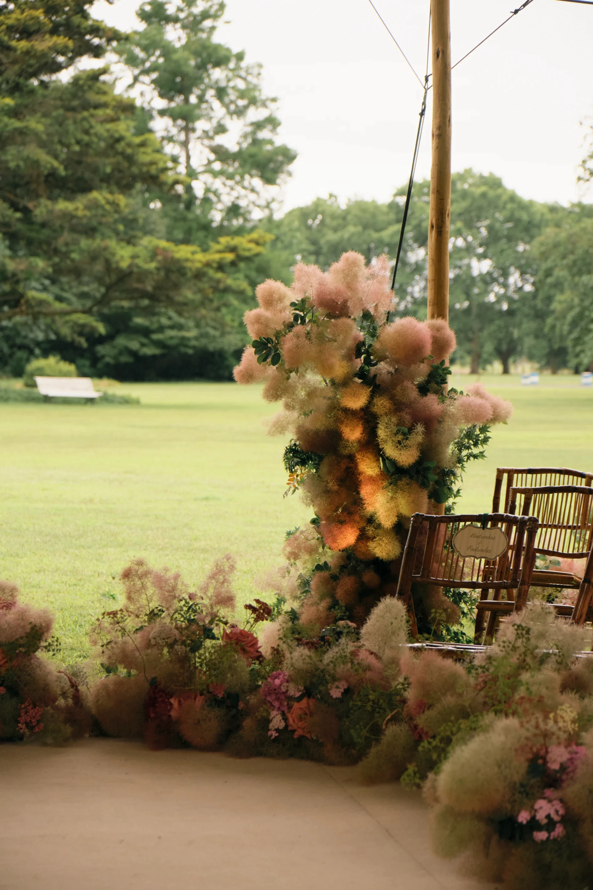 Estructura floral alta y vaporosa junto a sillas de madera bajo una carpa tensada para evento al aire libre.