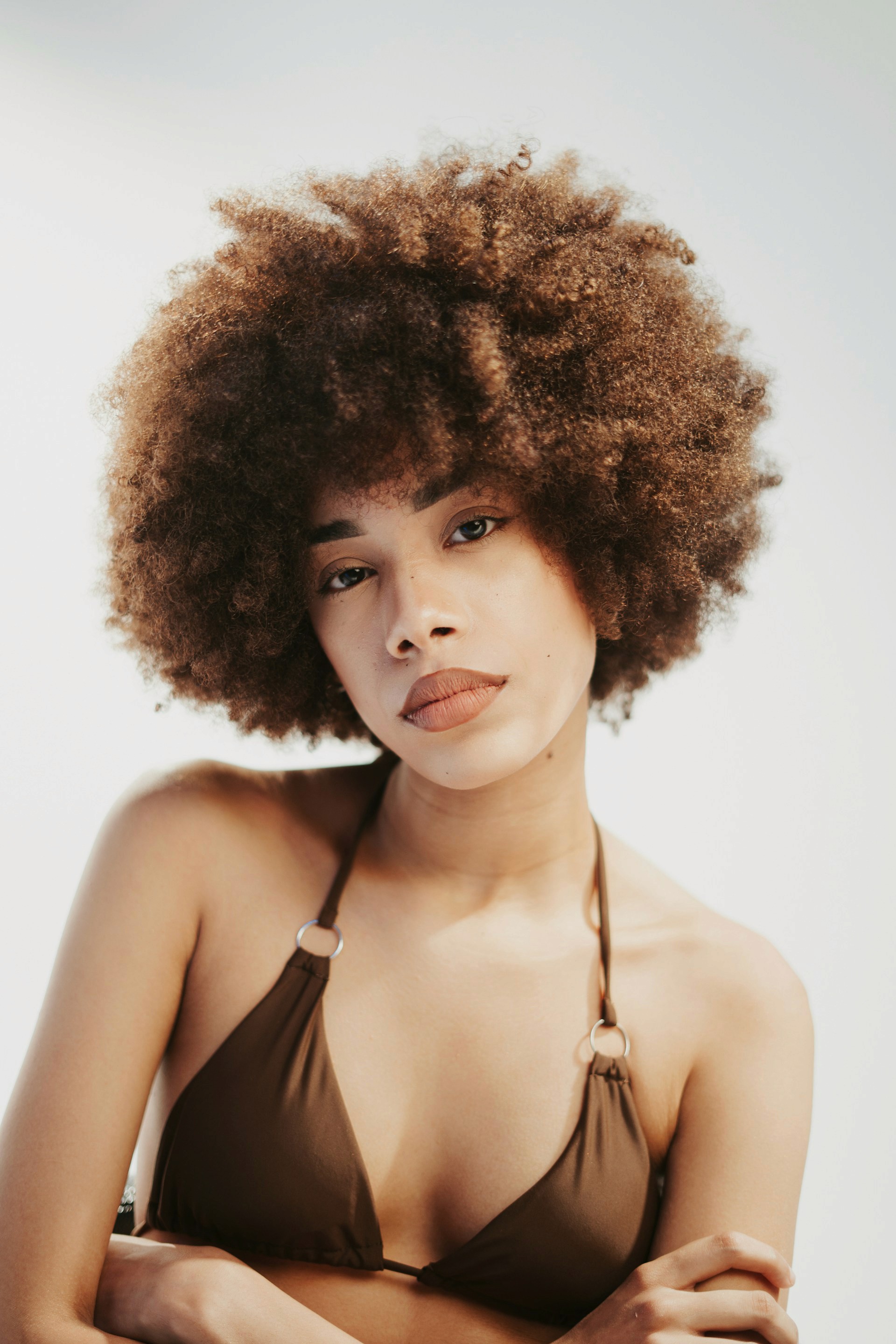 A woman with a large, voluminous brown afro posing in a brown bikini top against a bright, minimalist background.