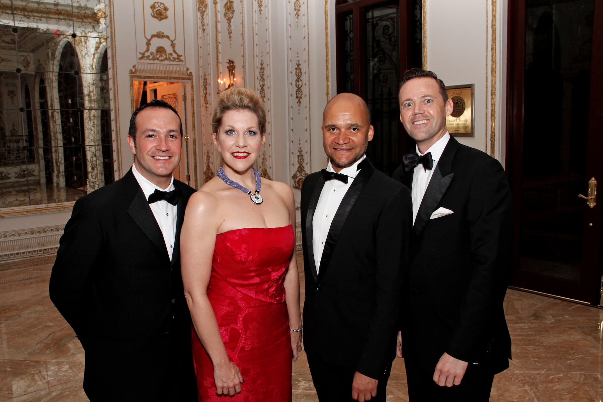 A woman in a red gown stands with three men in tuxedos inside a ballroom