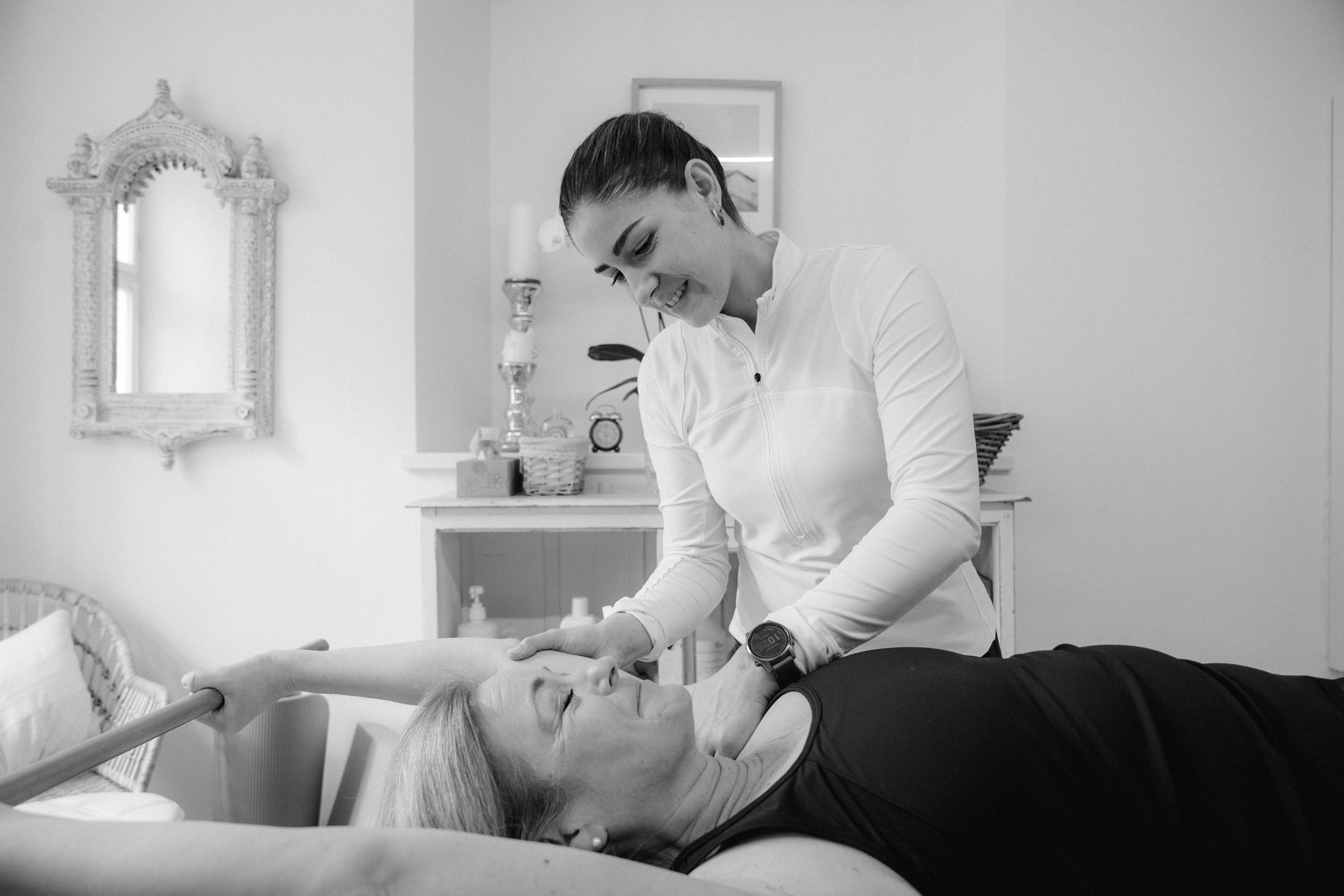 Therapeutin behandelt Patientin mit Physiotherapie. Frau liegt entspannt auf der Liege, während die Therapeutin den Nacken berührt.