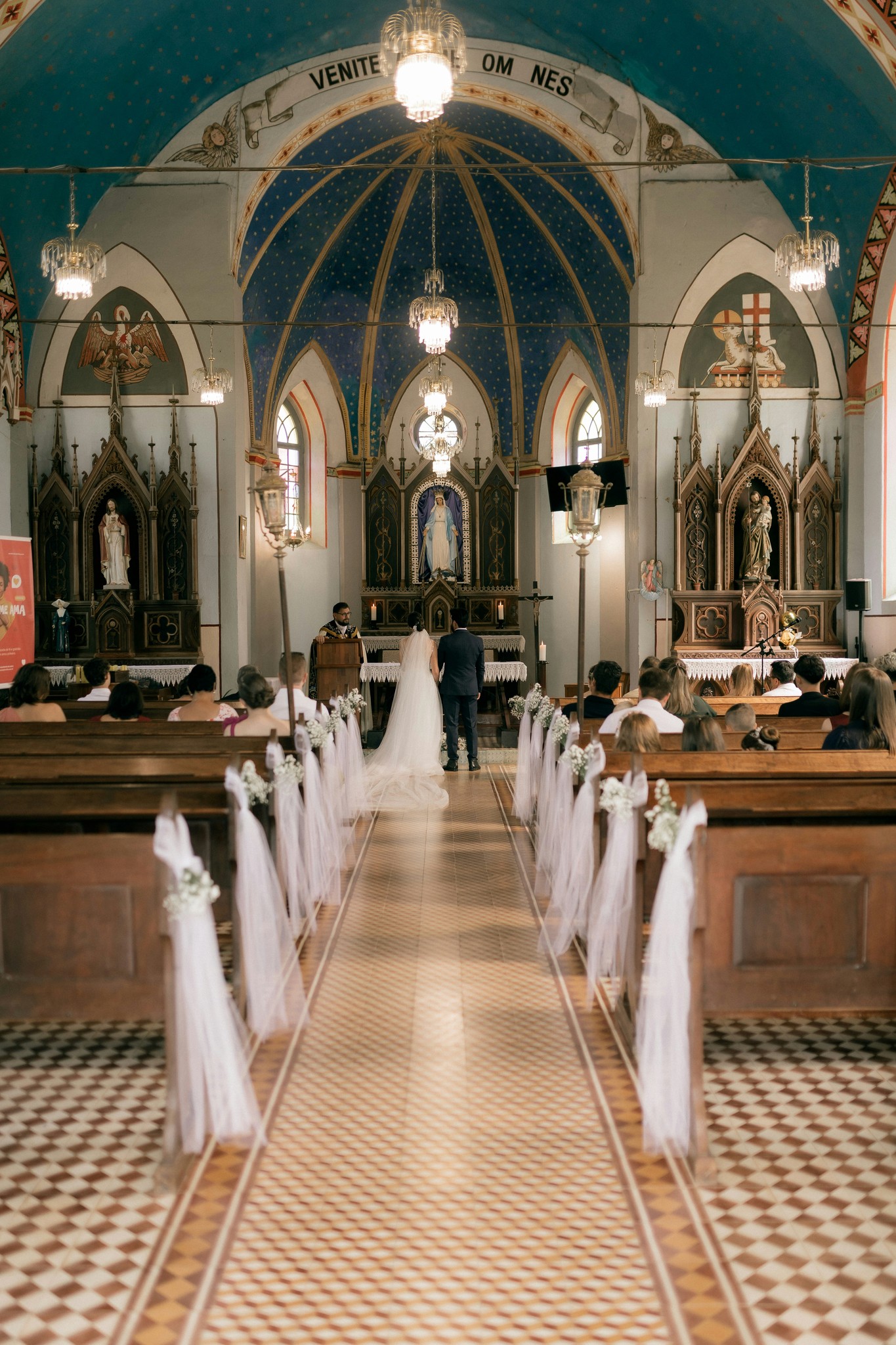 Bride and groom standing at the altar during a wedding ceremony inside an ornate church with blue vaulted ceilings and tulle-draped pews
