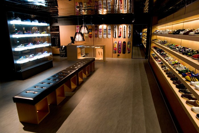 Wide view of the Apartment Store interior featuring illuminated sneaker walls, skateboard deck displays, a central leather bench, and warm timber joinery under low ambient lighting.