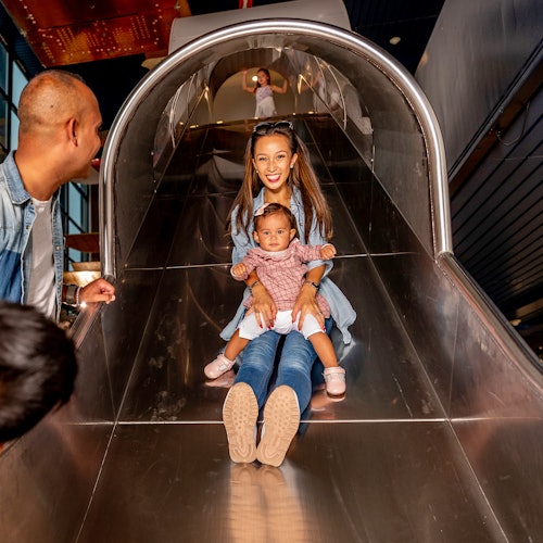 A woman and a toddler slide down a metallic playground slide. A man stands to the side, smiling at them.