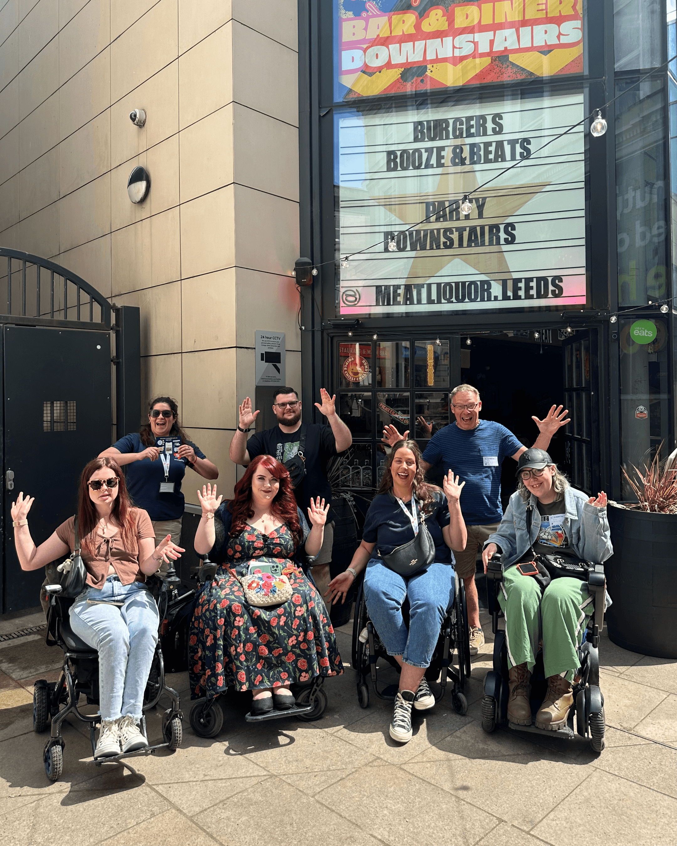 Our team celebrates inclusive places. Wheelchair users, and non-Disabled allies wave their hands in the air on a Sociability community day.