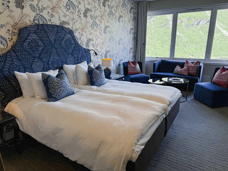 A beautifully designed hotel room features a large double bed with a patterned blue headboard, adorned with decorative pillows, set against a floral accent wall, and complemented by a window view of lush greenery, alongside two plush lounge chairs and a circular table.