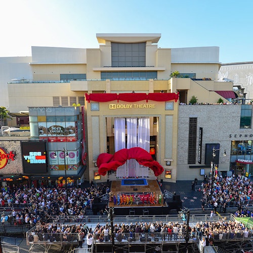 Crowd gathered outside Dolby Theatre, which is decorated with a large red ribbon, surrounded by shops and restaurants.