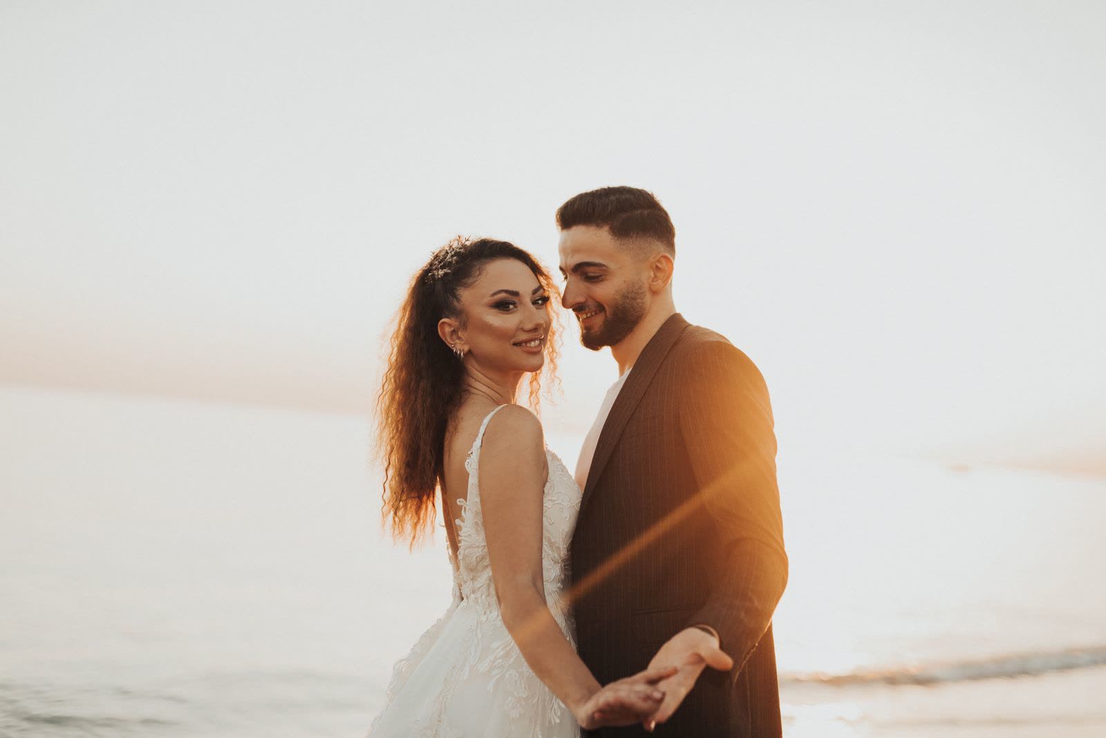 Couple in wedding clothes smiling on a beach with sunlight flaring behind them