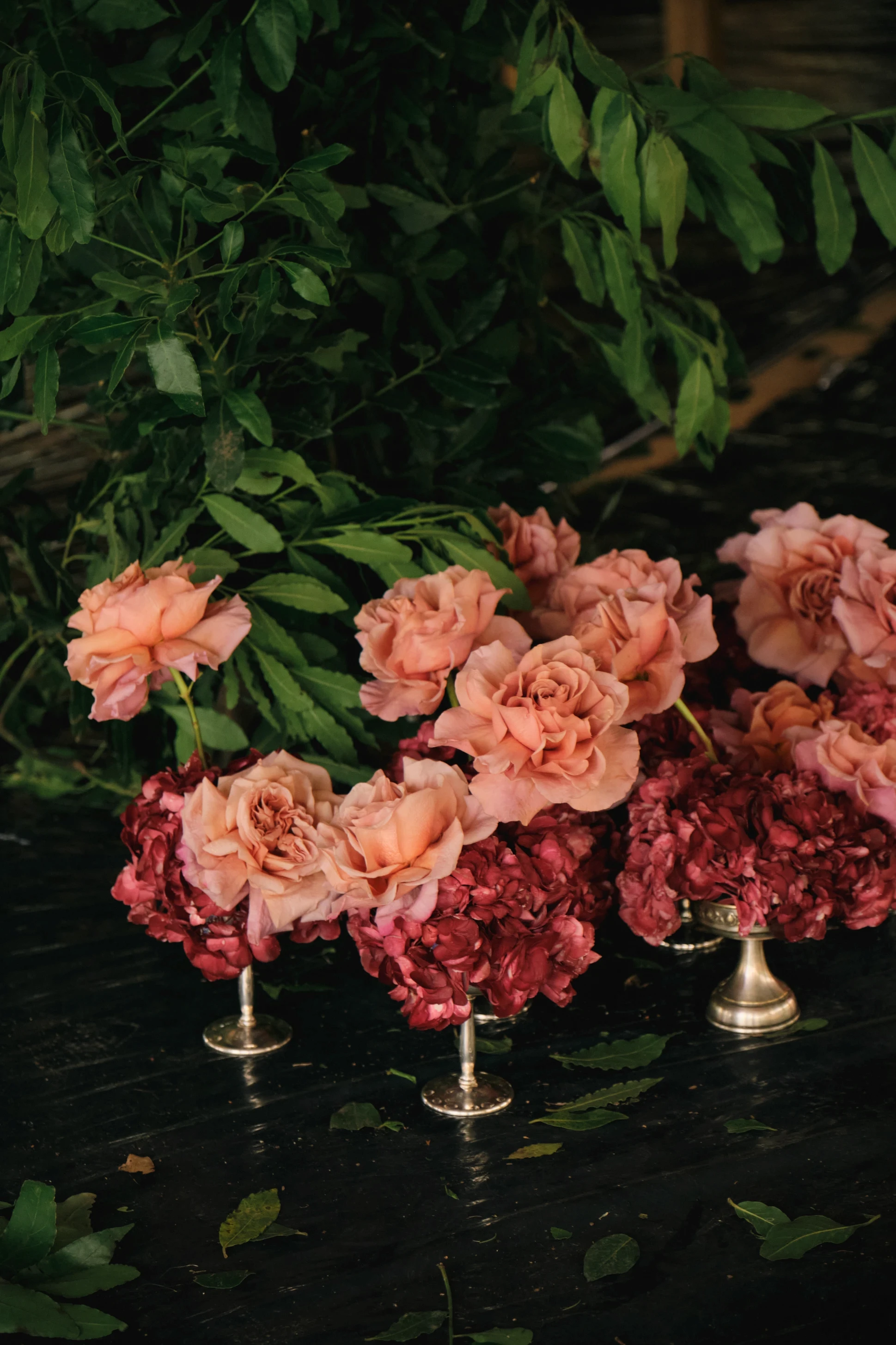 Fila de pequeños floreros plateados con rosas color durazno y hortensias bordó sobre una mesa oscura.