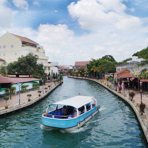 A blue and white boat cruises along a canal flanked by buildings, trees, and a pedestrian walkway under a partly cloudy sky.
