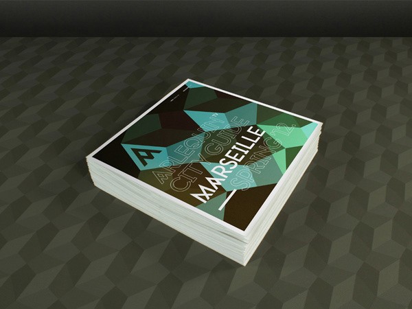 A stack of white cards rests on a dark gray surface with a geometric pattern. The top card displays “MARSEILLE” in white text against a black background with teal geometric shapes. A stylized “A” logo appears in the top left corner. The cards are neatly stacked and angled slightly.