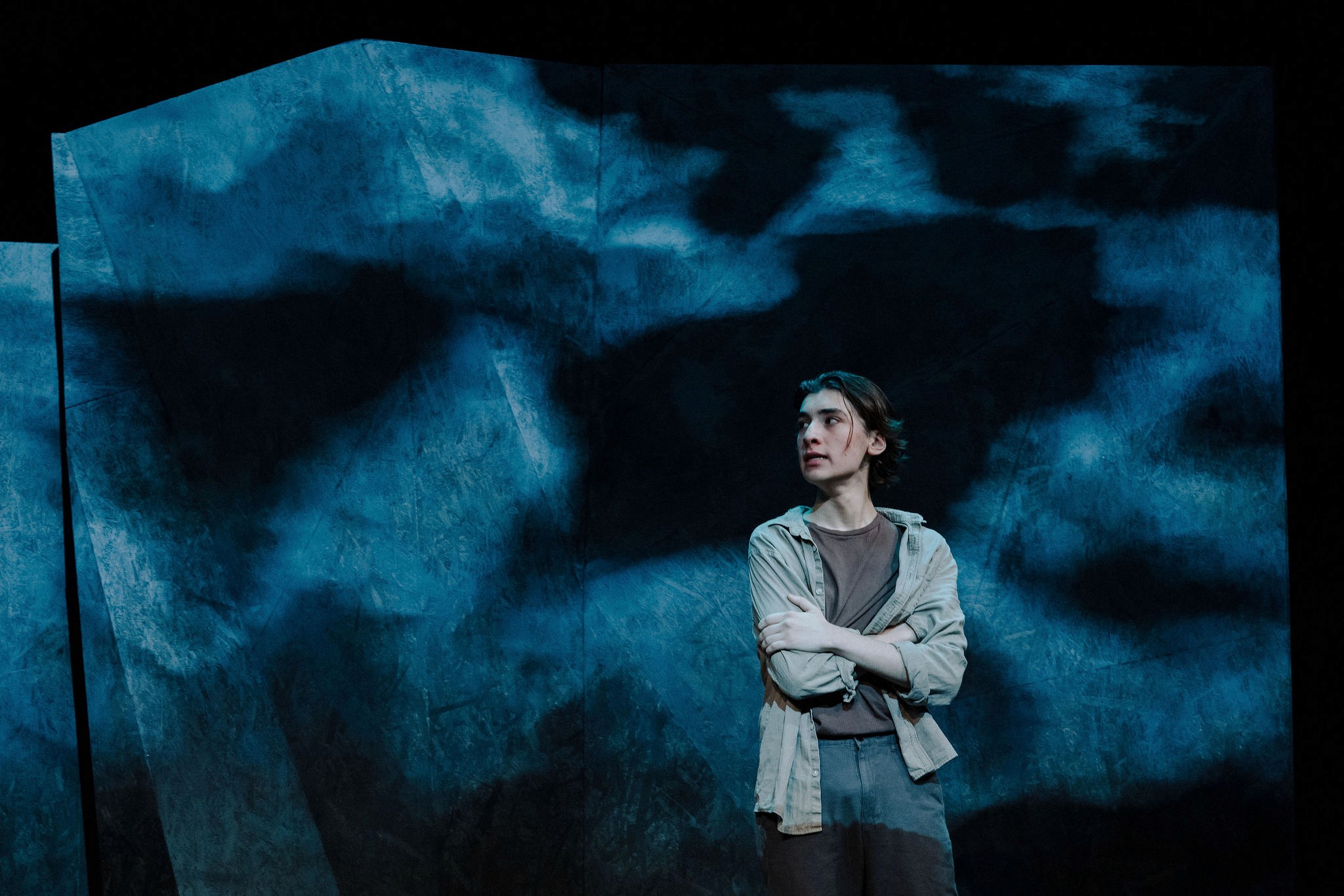 A performer stands alone with arms folded against a large textured backdrop resembling stormy sky or ocean.
