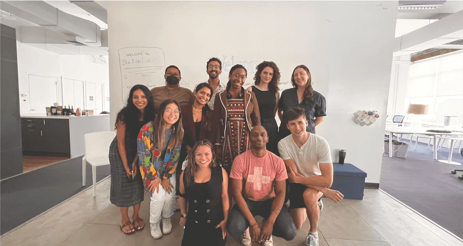 A group of diverse design, product, engineering and subject matter expert fellows stand in front of a whiteboard that says "Welcome to Blue Ridge Labs"