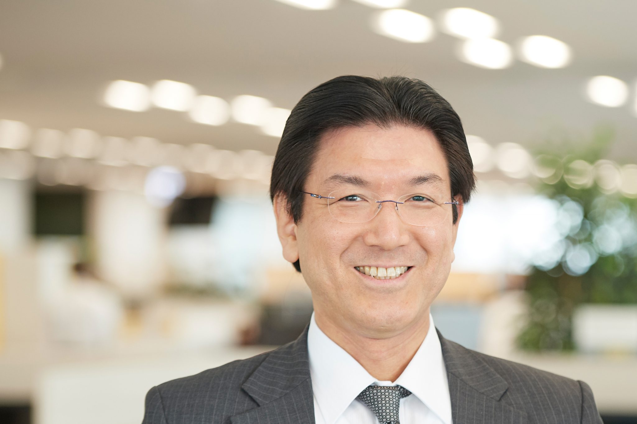 A smiling professional man in a suit, standing in a modern office environment with bright lighting.