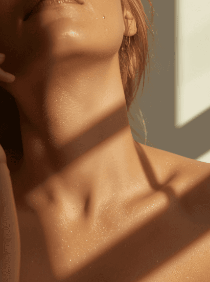 Close-up of a woman's neck and chin bathed in warm sunlight. Shadows create a serene, intimate atmosphere, highlighting soft skin texture.