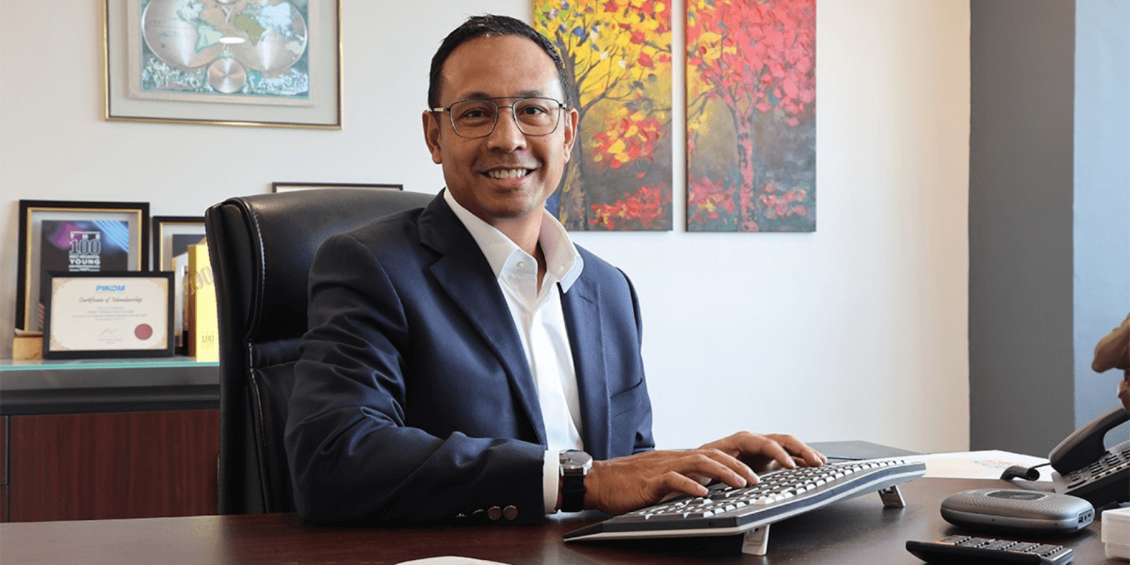 "Business professional smiling at his desk with certificates and artwork in the background, representing growth and success."