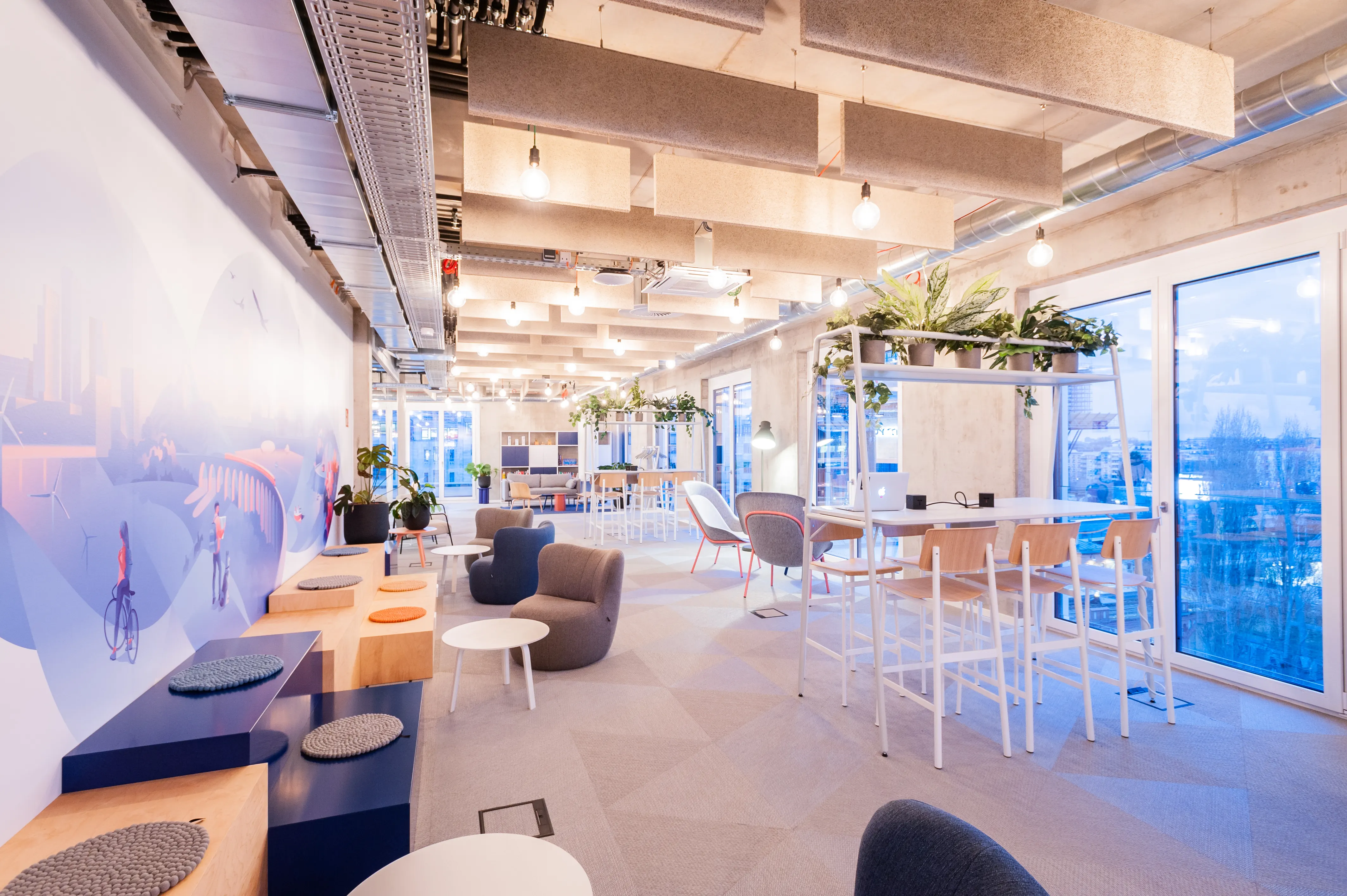 Work café at OMIO with a wall mural in soft blues, a wooden bench with felt seat pads, navy sideboards, grey armchairs, white standing tables with plant racks and floor-to-ceiling windows.