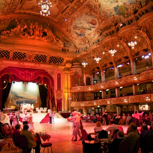 People dancing in an ornately decorated ballroom with a large stage, red curtains, and detailed ceiling artwork.