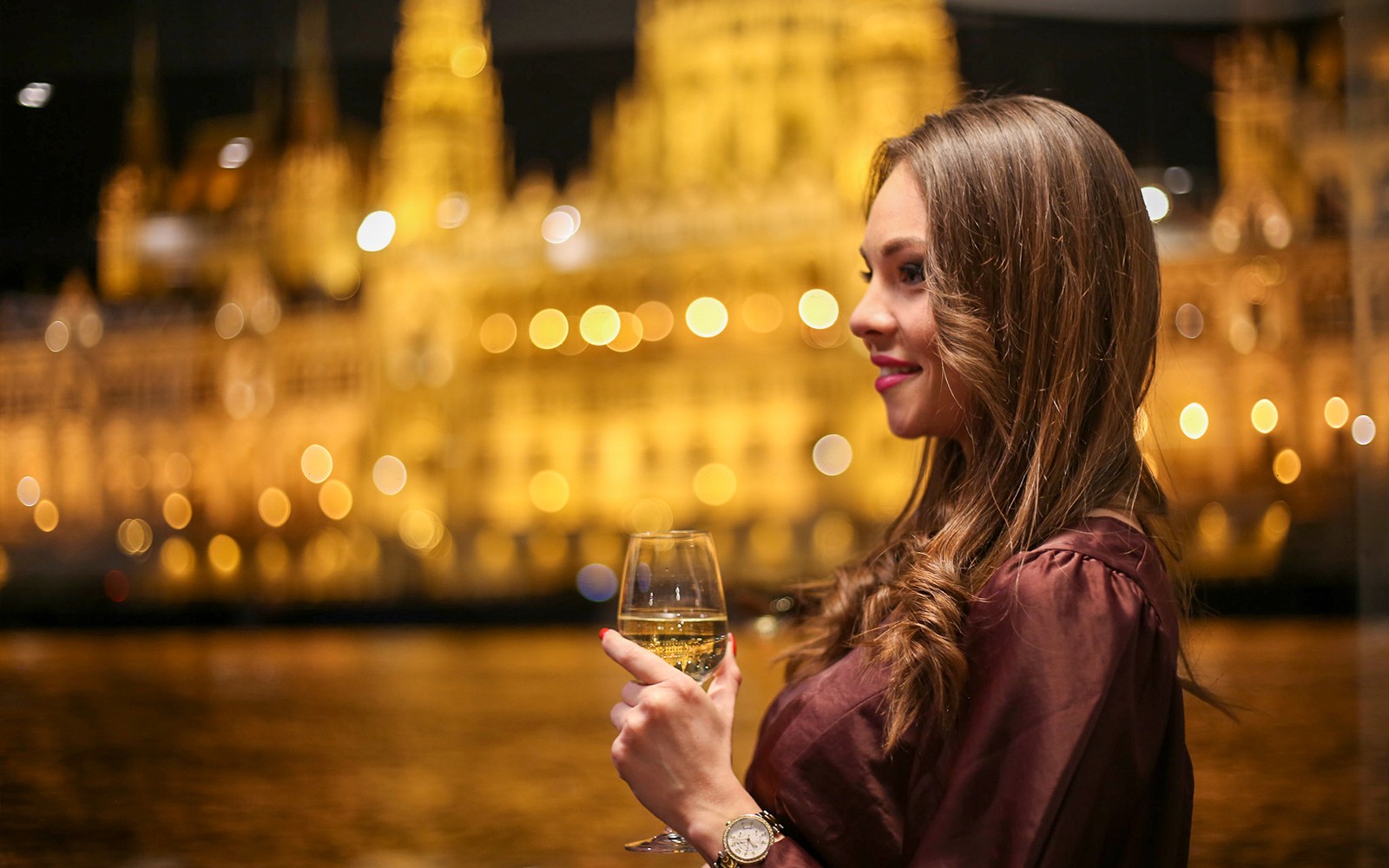 Woman enjoying a drink on Budapest dinner cruise with illuminated cityscape.