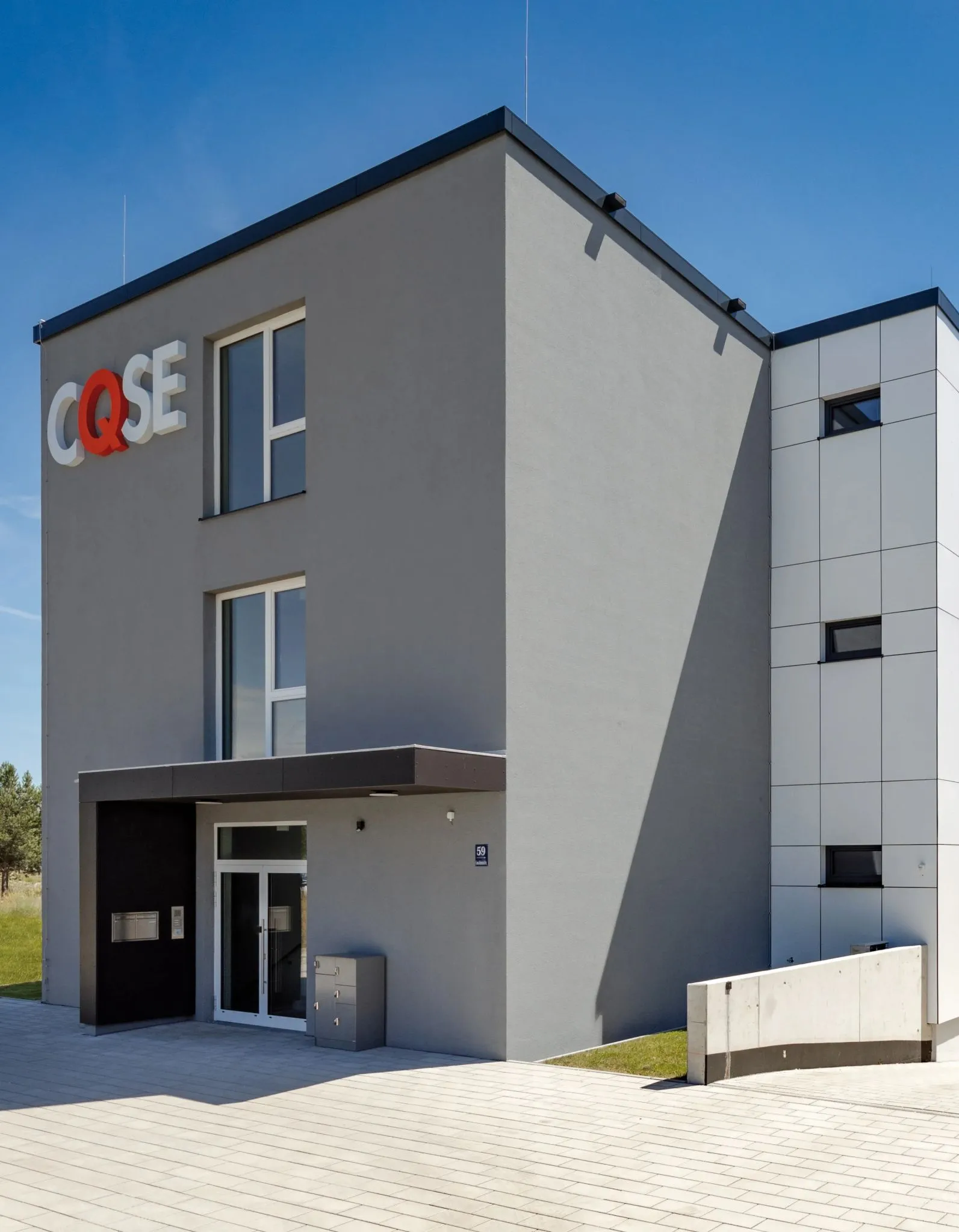 Modernes Bürogebäude mit Eingangsbereich, Fenster und dem Schriftzug "CQSE" in Rot und Weiß an der grauen Fassade.