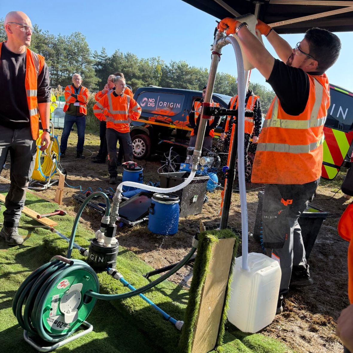 A worker in orange safety gear pours water from a large plastic jug into an apparatus under a tent, surrounded by other workers and equipment.