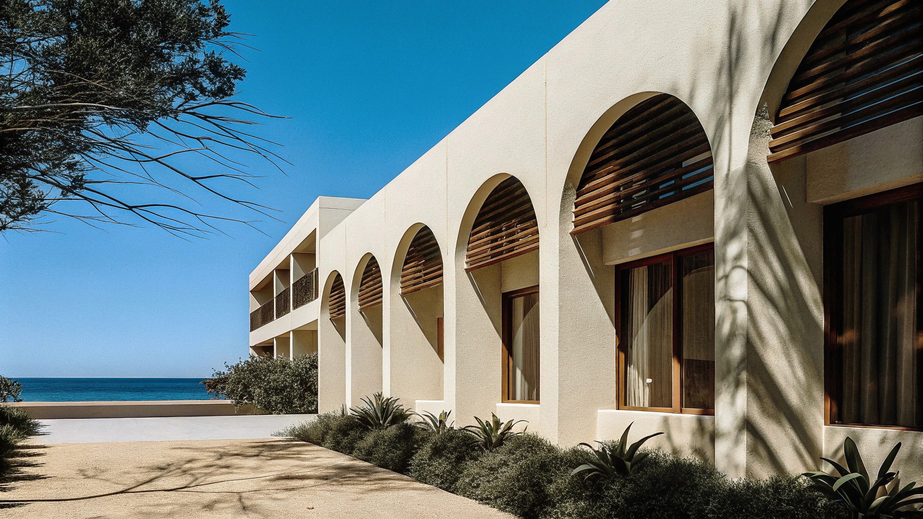 Sun-drenched frontal shot of a modern Mediterranean hotel with arched wooden windows, minimal landscaping, smooth white architecture, calm ocean in the background, cinematic tone.