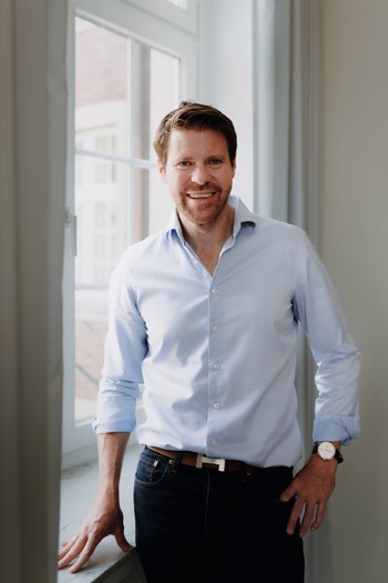 Jüngerer männlicher Geschäftsführer im hellen Hemd steht entspannt vor einem Fenster im Büro von SLB.