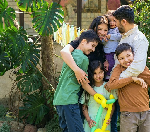 A large, happy family with a mother, father, and four children hugging outdoors near tropical plants and party decorations.