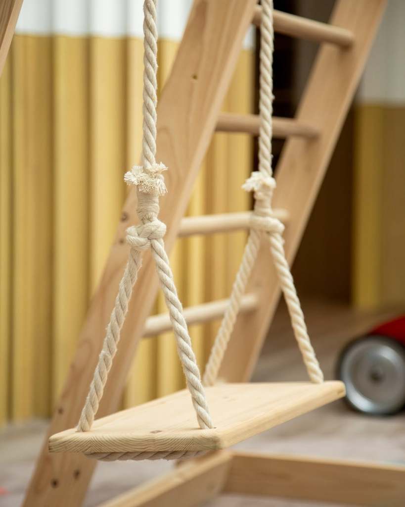 A wooden swing hanging from a ladder made of natural wood, with braided rope supports and a simple, rectangular seat.