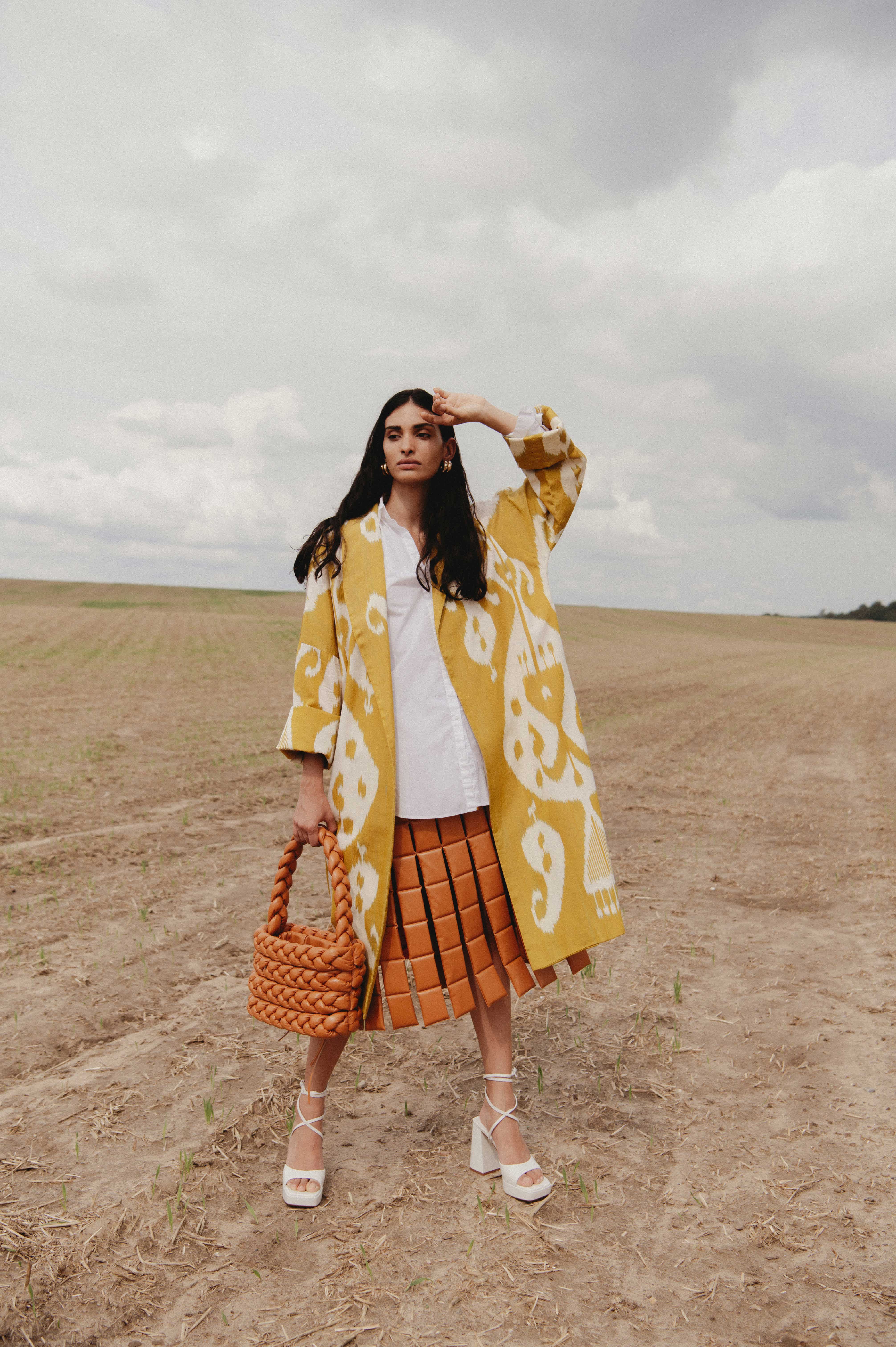 Woman standing in a field wearing a white shirt and long yellow patterned coat, shielding her eyes from the sun.