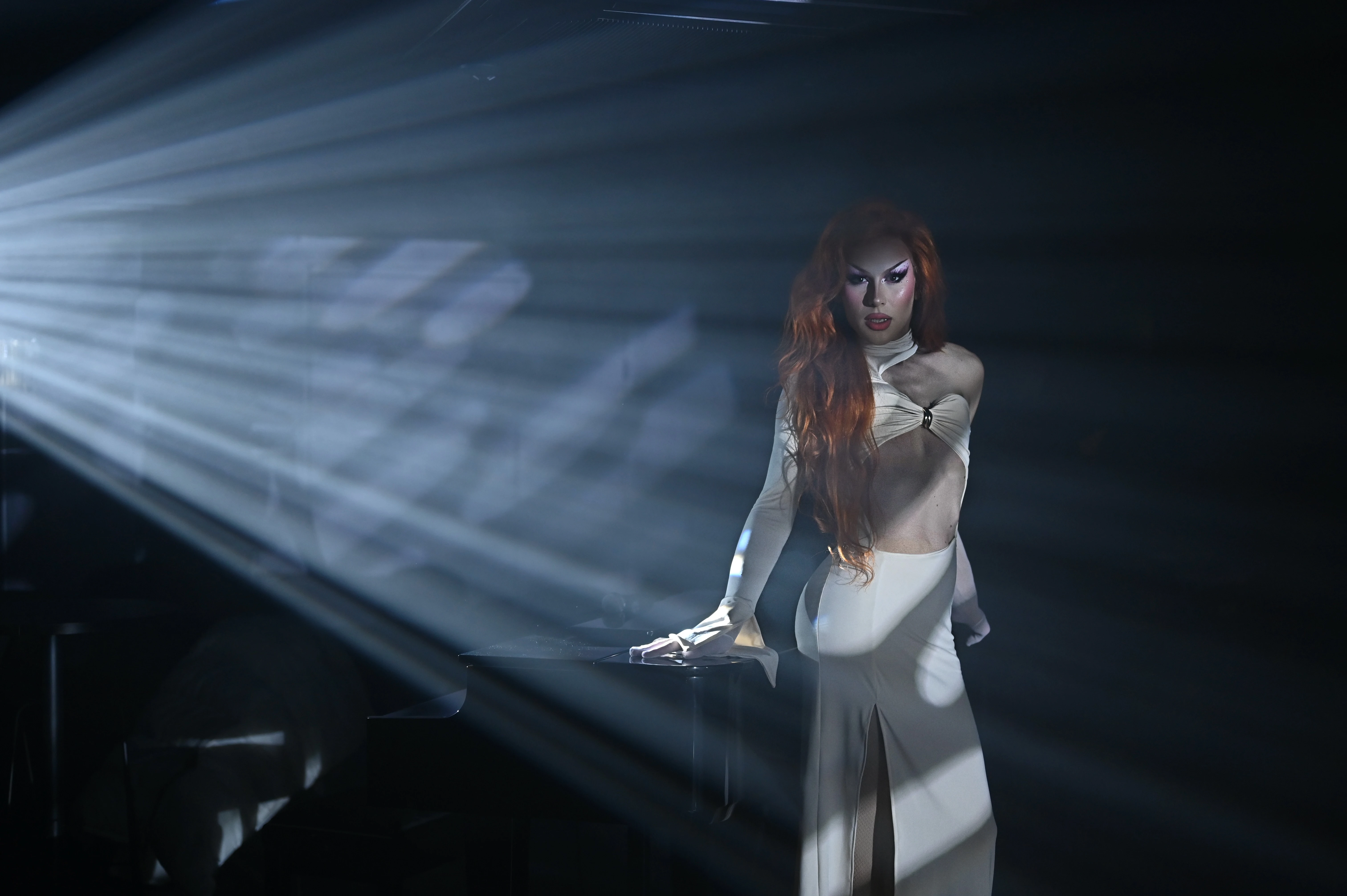 A glamorous cabaret performer with long hair, wearing an elegant white cutout dress, standing mysteriously by a dark piano under dramatic, cinematic slatted lighting