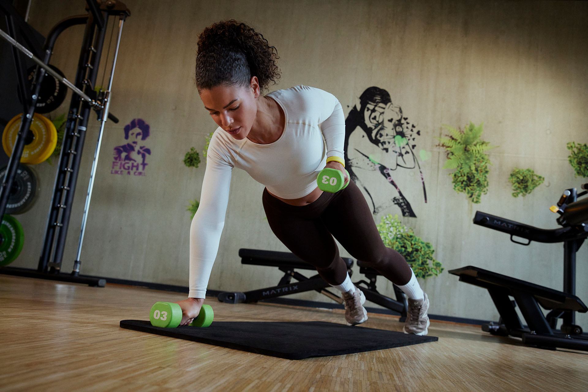 Eine Frau trainiert im modernen Lady-Gym von XTRAFIT. Sie hält die Plank auf einer schwarzen Matte mit Kurzhanteln. Sie trägt ein weißes langärmliges Oberteil und braune Leggings. In ihrer linken Hand hält sie eine grüne Kurzhantel. Im Hintergrund sind Fitnessgeräte und eine Wandtapete mit Pflanzen zu sehen. Die Frau ist konzentriert und engagiert bei ihrem Training.