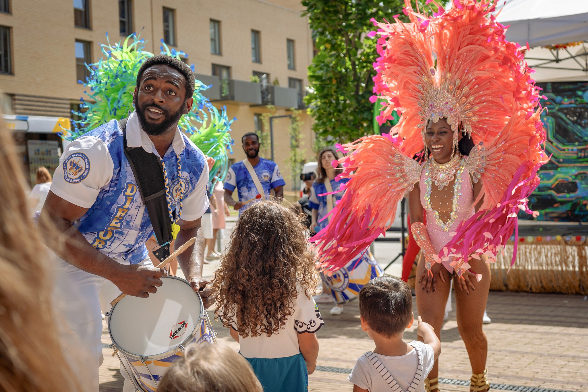 Carnival performers dance and play drums outdoors, wearing bright blue, green, and pink feathered costumes, as smiling children watch and interact with them in a sunlit urban square.