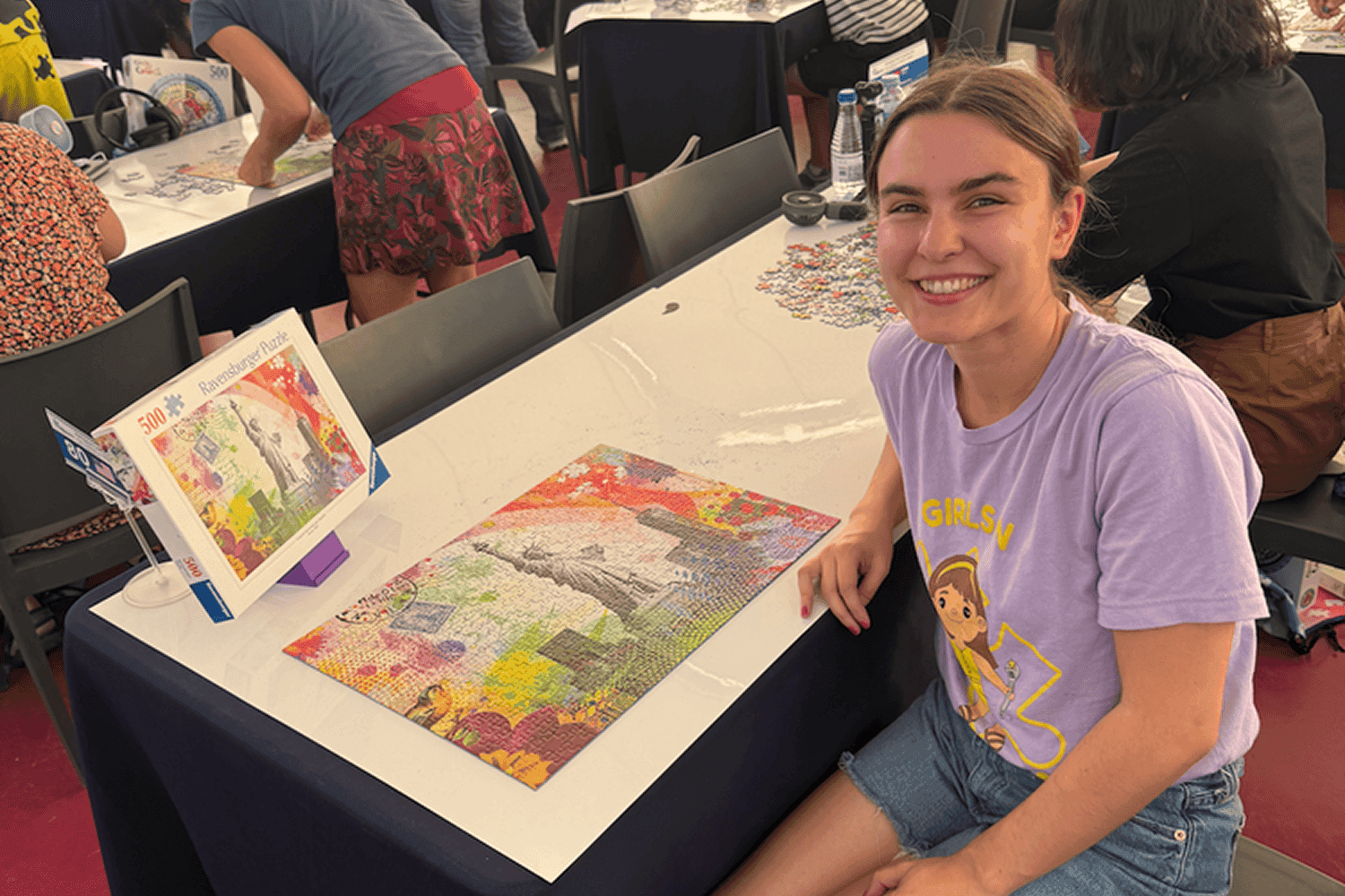 Uma pessoa vestindo uma camiseta roxa está sentada, sorrindo, ao lado de um quebra-cabeça colorido concluído em uma mesa, em uma sala movimentada com outras pessoas montando quebra-cabeças por perto.