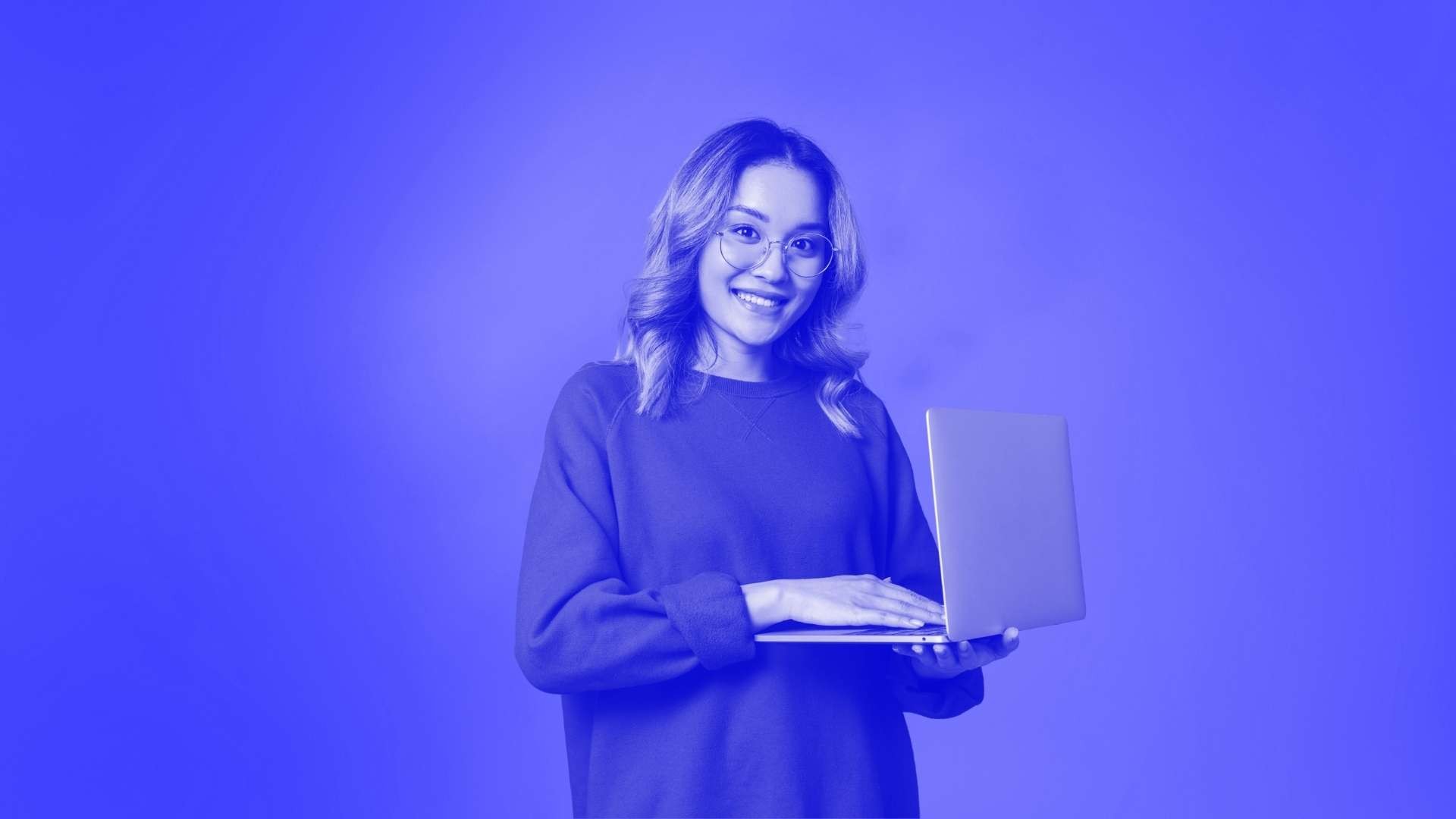 A woman wearing glasses and a jumper standing holding her laptop and typing