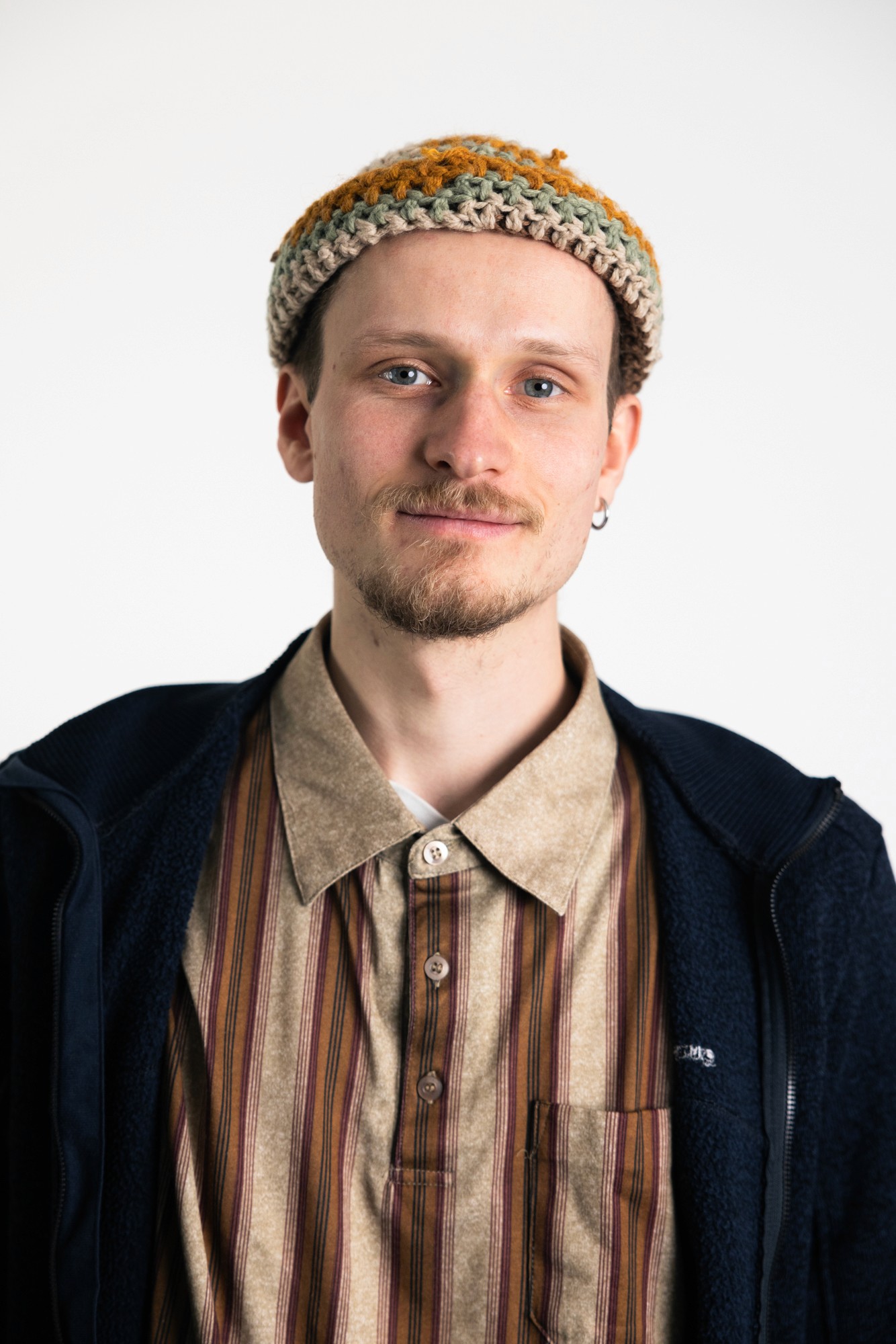 A young adult with a neutral expression, wearing a collared shirt and a hat, set against a plain background.