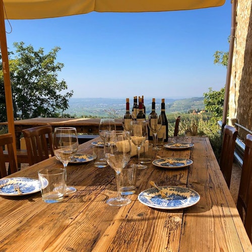 Mesa rústica al aire libre con copas de vino vacías, platos con migas y varias botellas de vino. Vista panorámica del campo en el fondo.