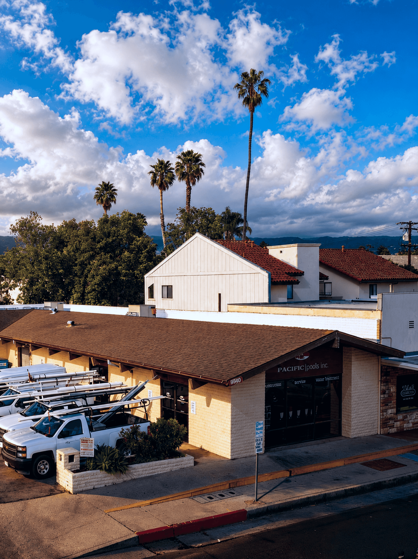 Aerial view of a single-story commercial strip building with a brown hip roof, covered entry canopy, and a surface parking lot with service vehicles, surrounded by mature palm trees and residential neighborhood in a Southern California coastal community.